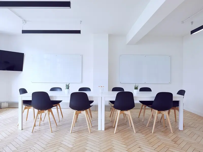 An empty white meeting room with a long table, dark chairs and two glass boards