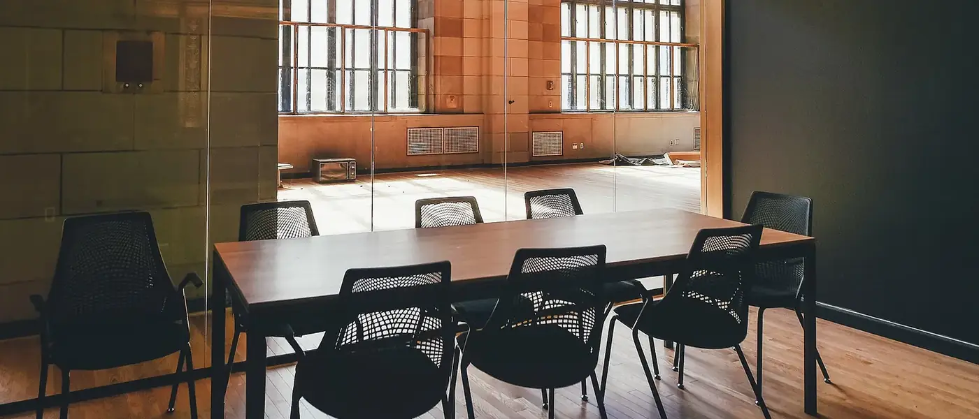 A meeting room with a dark wooden table and mesh chairs beside tall factory windows