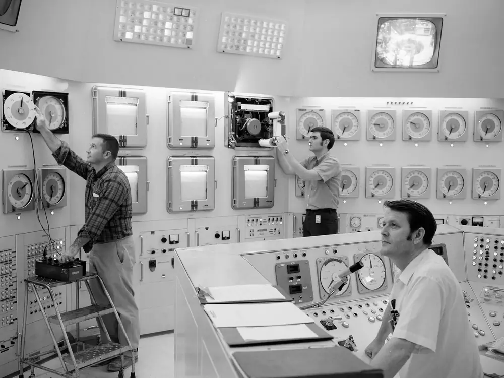 Three technicians reading a wall of analogue dials and chart recorders in a control room