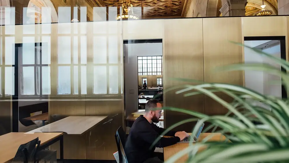 A person working at a laptop in an open office seen through a glass partition